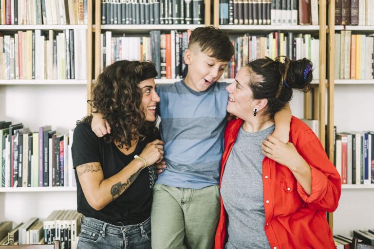 Two women standing in front of a bookcase with a young boy in the middle with his arms wrapped around both of them.