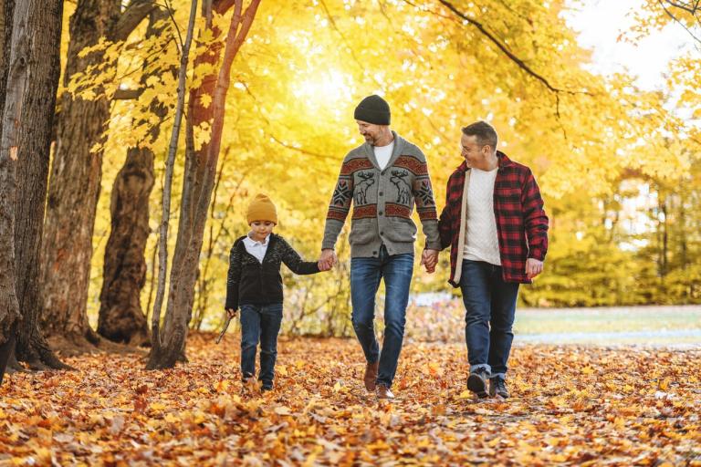 A happy two-man couple walking through a park in Autumn with a child holding one of the man's hand.