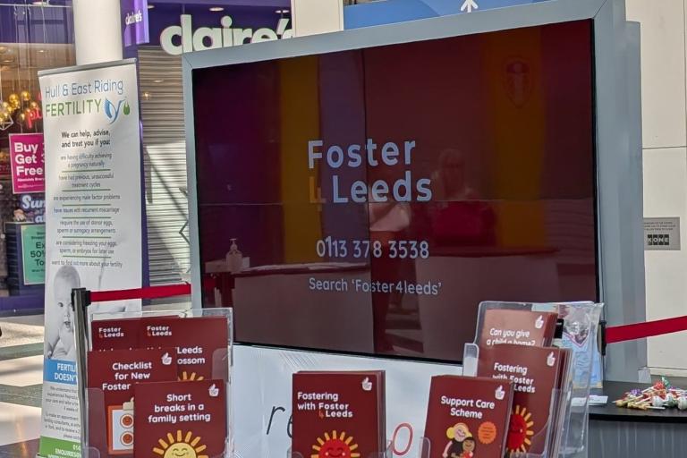 A stand with Foster4Leeds leaflets and booklets. Behind is a large television screen with the Foster4Leeds logo on it.