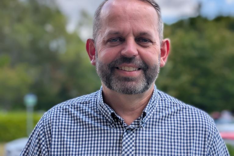 Rob poses for a photo, smiling. He is wearing a blue chequered, short-sleeve shirt.