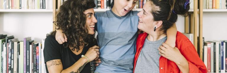 Two women standing in front of a bookcase with a young boy in the middle with his arms wrapped around both of them.