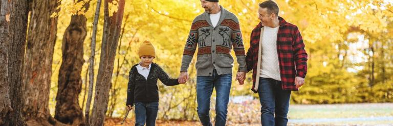 A happy two-man couple walking through a park in Autumn with a child holding one of the man's hand.
